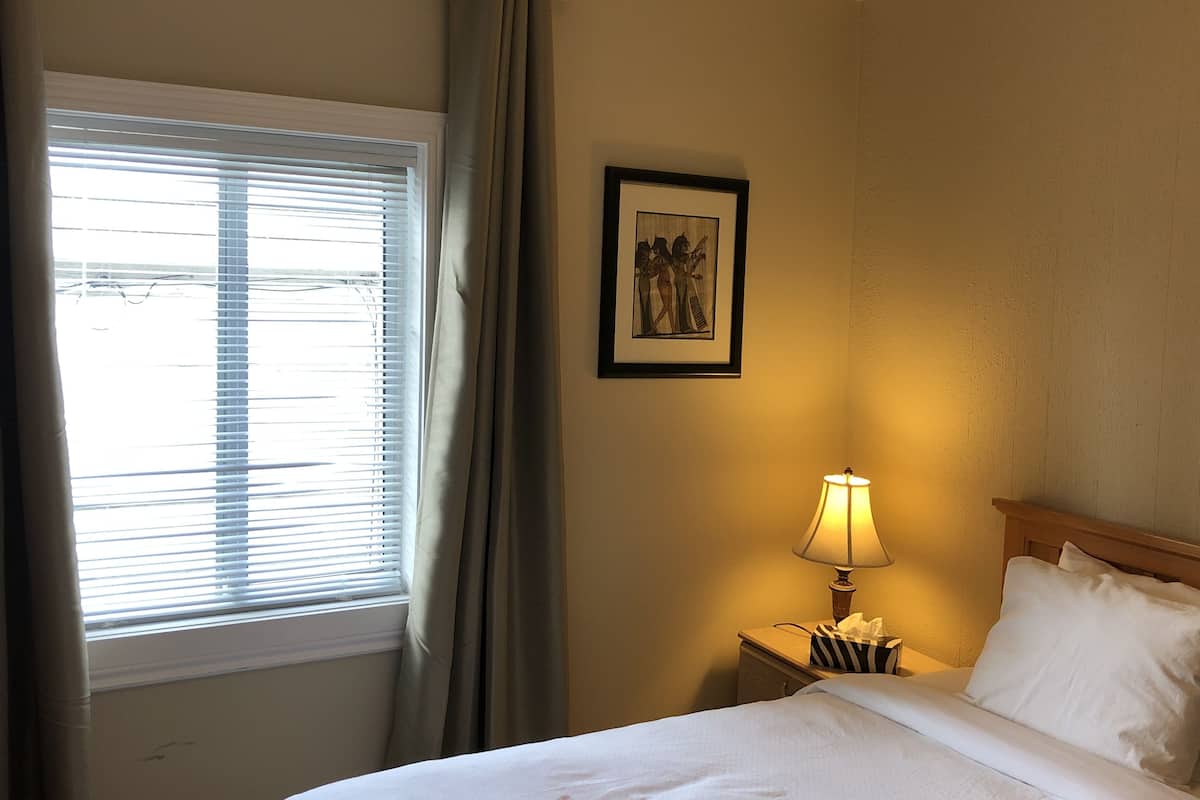 Una habitación de hotel con una cama, una mesita de noche con lámpara, un cuadro enmarcado en la pared y una ventana con cortinas.