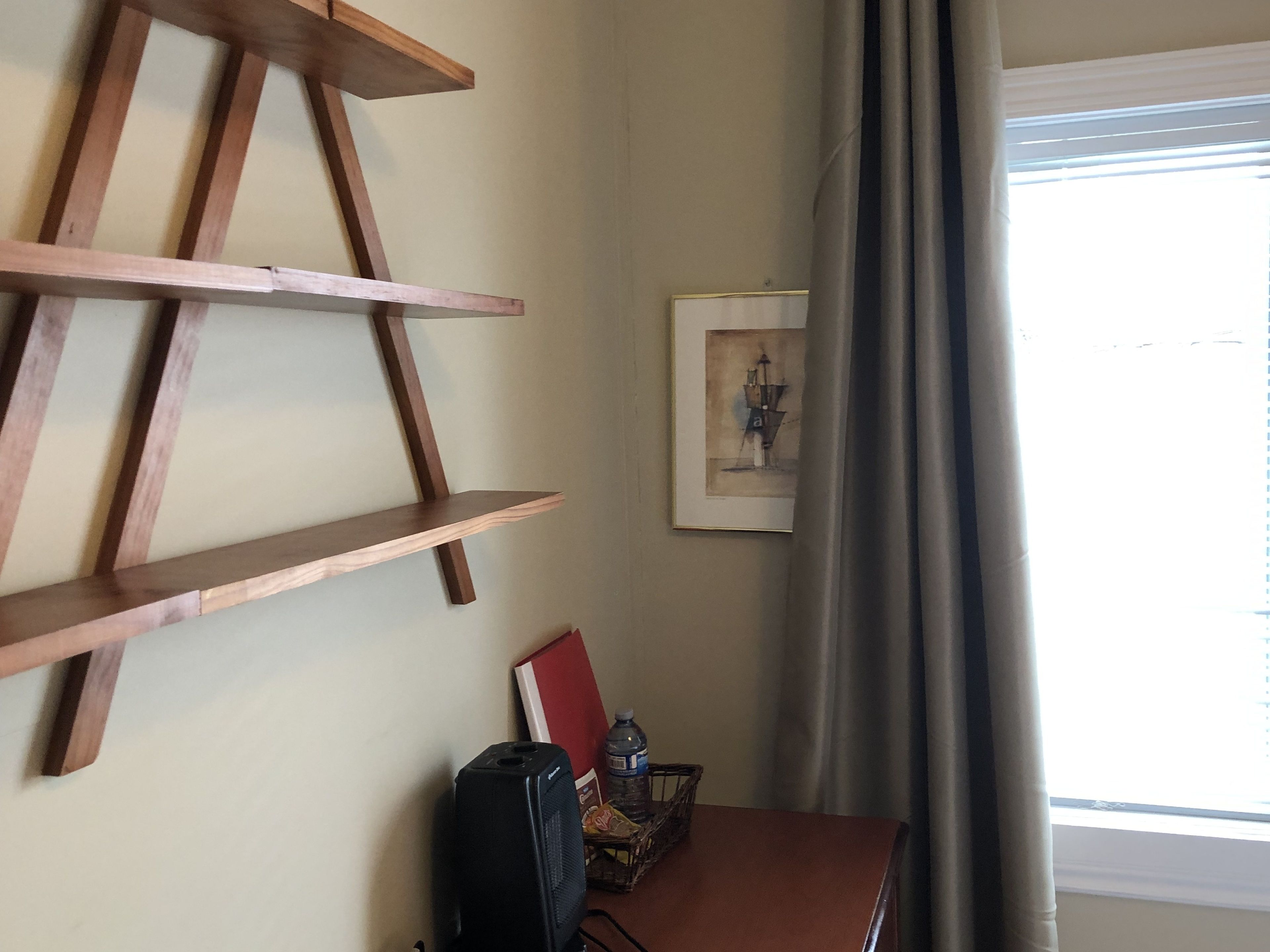 Una habitación con una estantería de madera escalonada, un cuadro enmarcado en la pared, una mesa con una maleta y una ventana con cortinas.