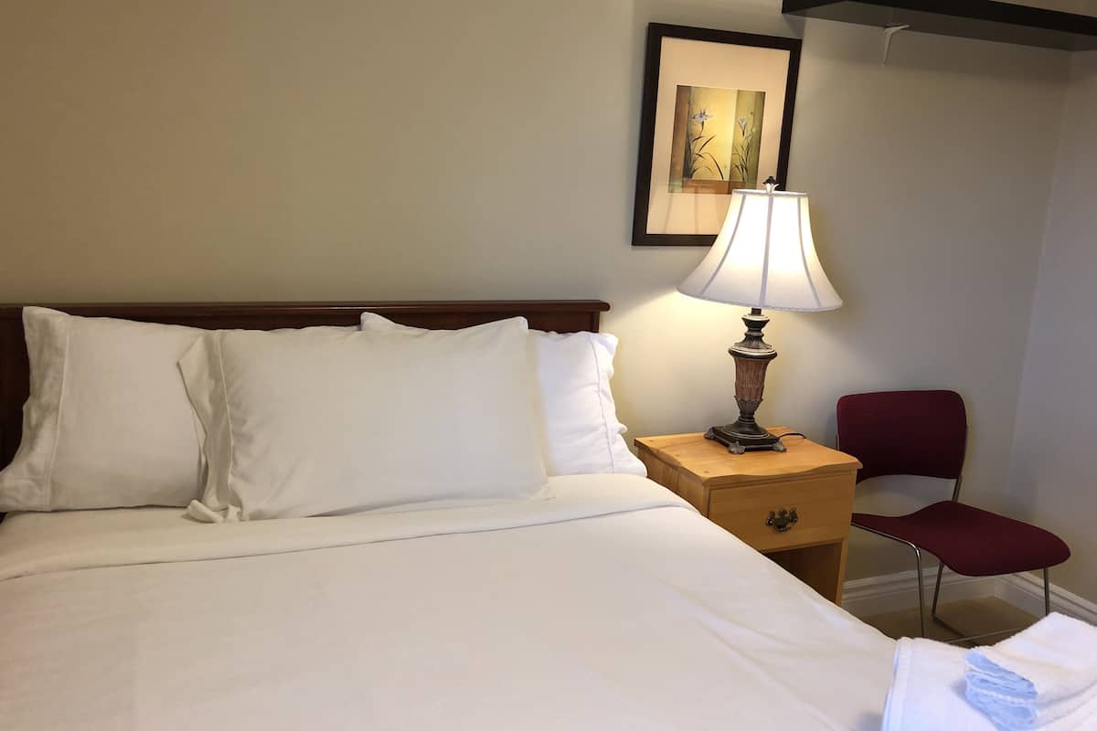 Una cama bien hecha con sábanas blancas, una mesita de noche de madera con una lámpara, una silla roja y un cuadro en la pared.