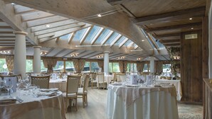 A spacious dining area with multiple round tables, wooden beams, and large windows allowing natural light. - Family & Wellness Hotel Shandranj (Tesero)