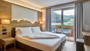 A hotel room with a large bed, wooden headboard, two bedside lamps, a view of a balcony with outdoor furniture, and a wooden floor. - Family & Wellness Hotel Shandranj (Tesero)