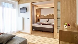 A modern hotel room with a wooden floor, a bed with a striped blanket, a TV mounted on the wall, and a wooden wardrobe with fruit on top. - Family & Wellness Hotel Shandranj (Tesero)
