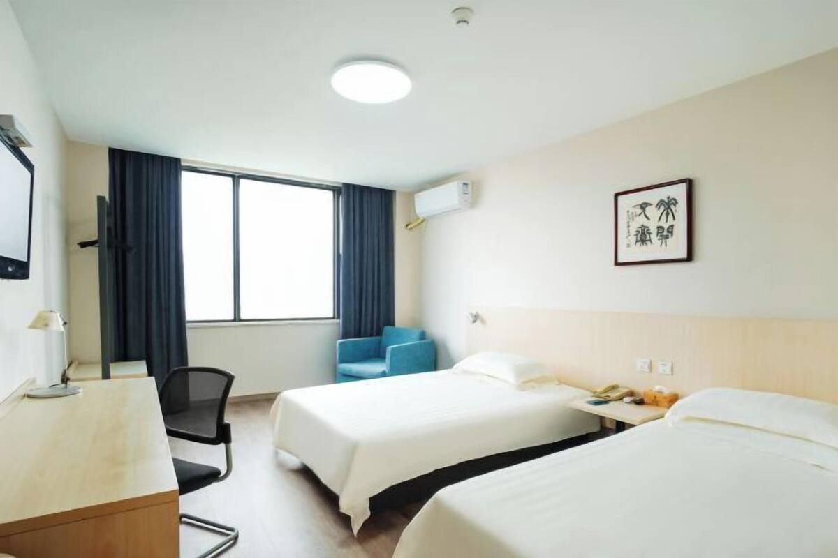 Habitación de hotel con dos camas, un escritorio, una silla, una ventana con cortinas, un equipo de aire acondicionado y un cuadro en la pared.