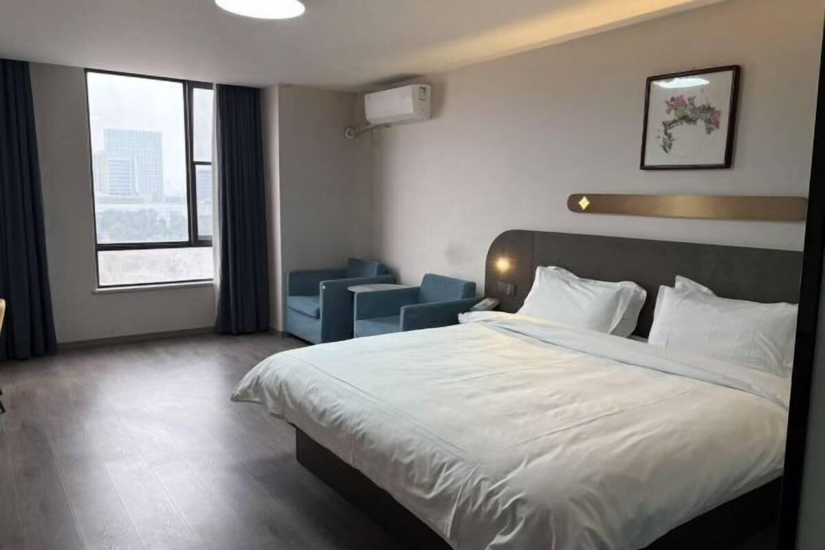 Habitación de hotel con una cama grande, una zona de estar con sillas, una ventana con cortinas y un equipo de aire acondicionado en la pared.