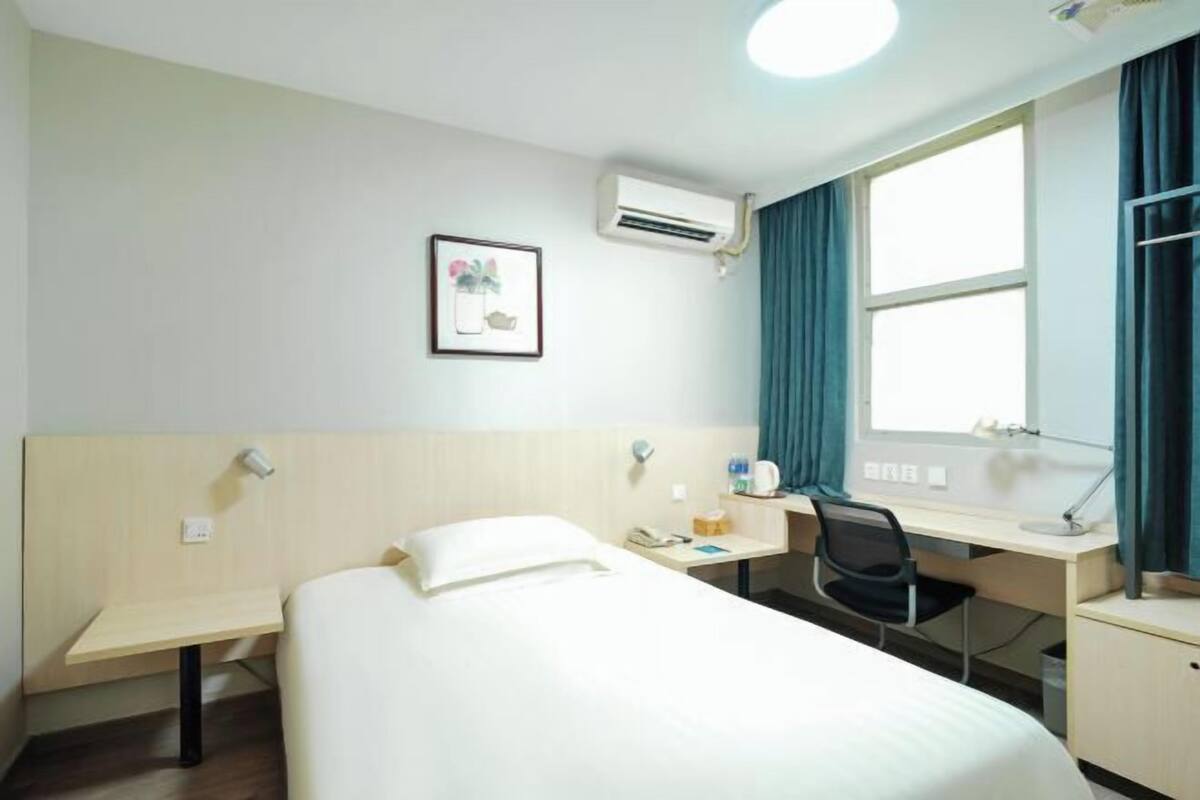 Habitación de hotel con cama, escritorio, silla, aire acondicionado, ventana con cortinas e imagen en un marco en la pared.