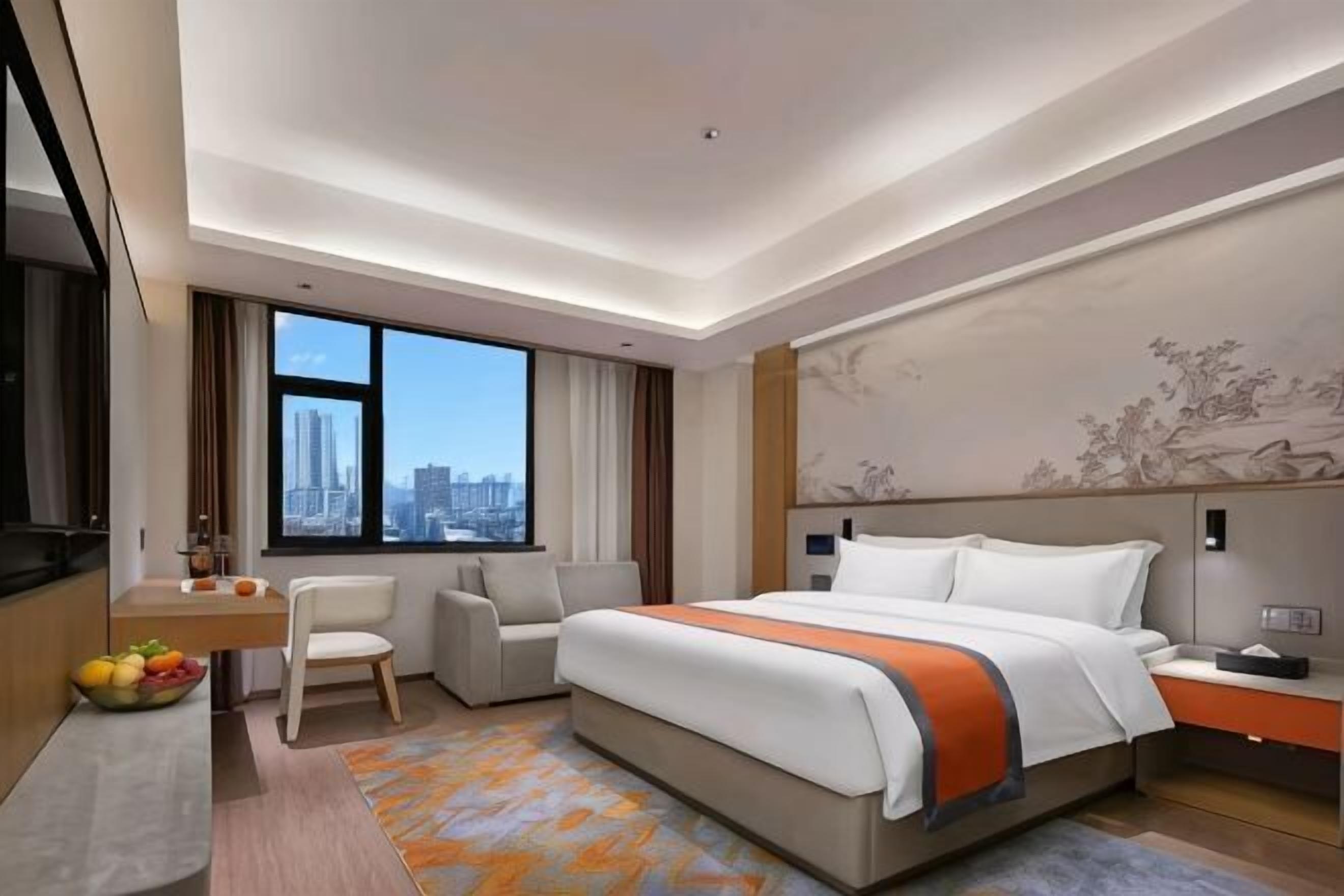Habitación de hotel con cama, escritorio, silla, televisor y una ventana grande con vista a la ciudad.