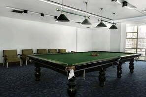Una mesa de billar en una habitación con alfombra estampada, sillas e iluminación colgante.