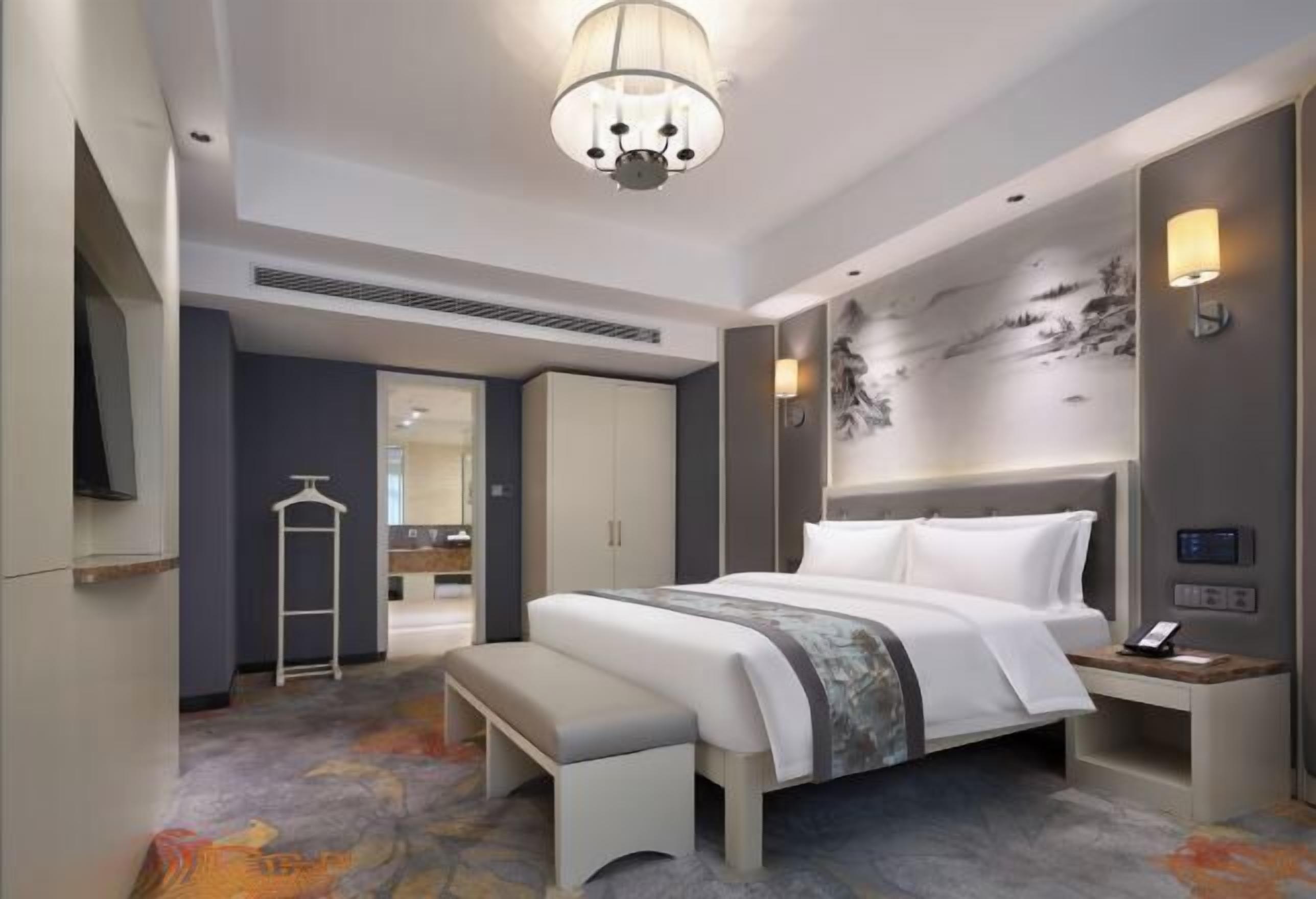 Quarto de hotel moderno com uma cama grande, mesa de cabeceira, banco, guarda-roupa e uma televisão.