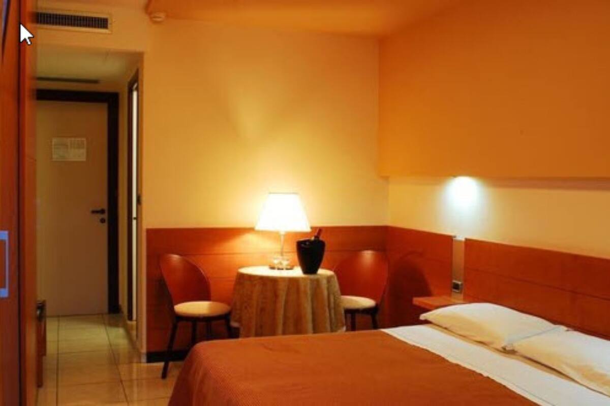 A hotel room with a bed, a table with a lamp, two chairs, and a door.