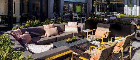An outdoor seating area with a sofa, chairs, and a small table, surrounded by plants and a building with a sign reading "PIVOT BAR X BALCONY".