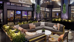 Un bar moderno y un área de balcón con un sofá grande, mesas y sillas, rodeado de vegetación e iluminado con luz ambiental.