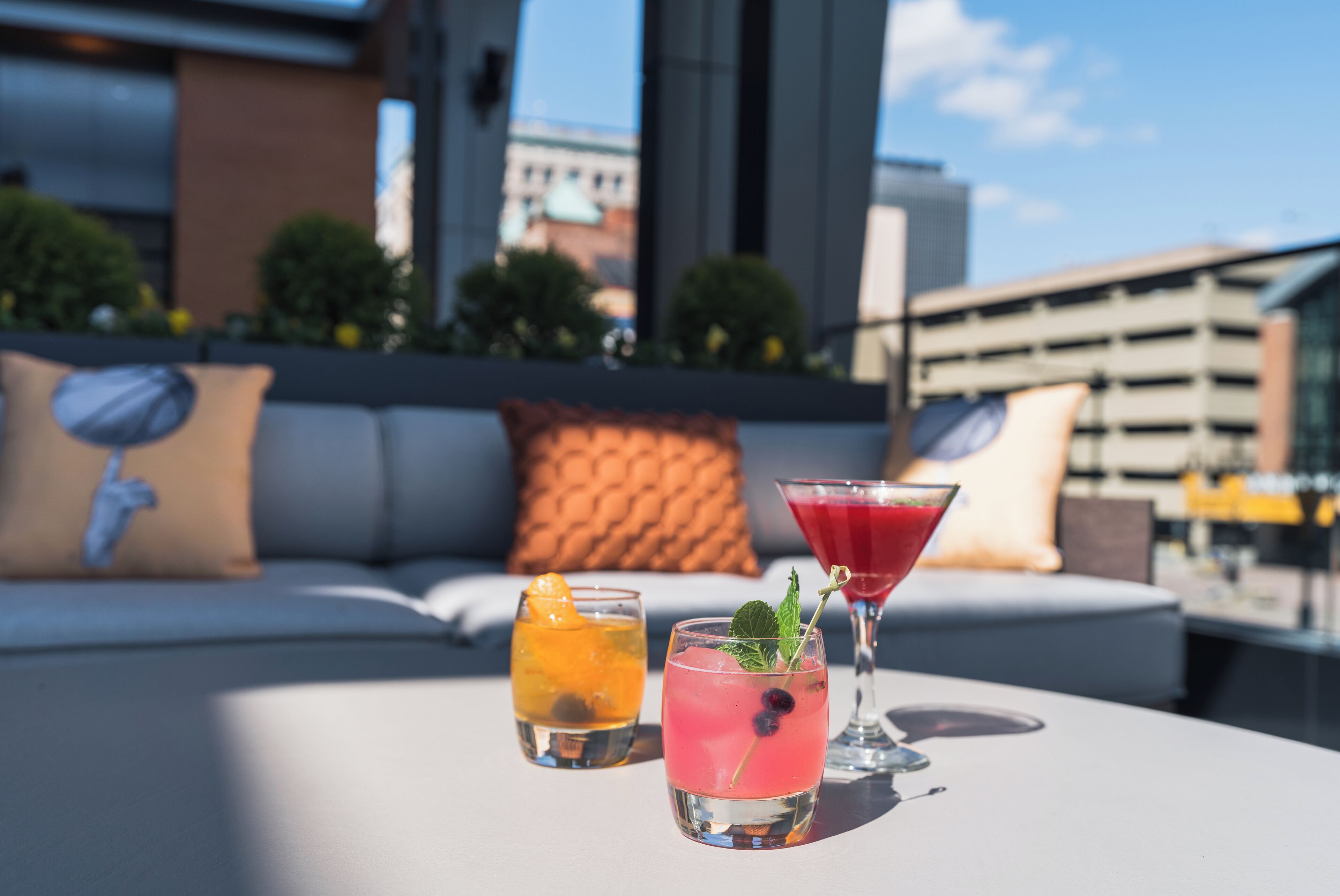 A rooftop terrace with two cocktails on a table, a sofa with cushions, and a cityscape view.