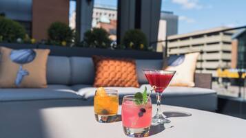 A rooftop terrace with two cocktails on a table, a sofa with cushions, and a cityscape view.