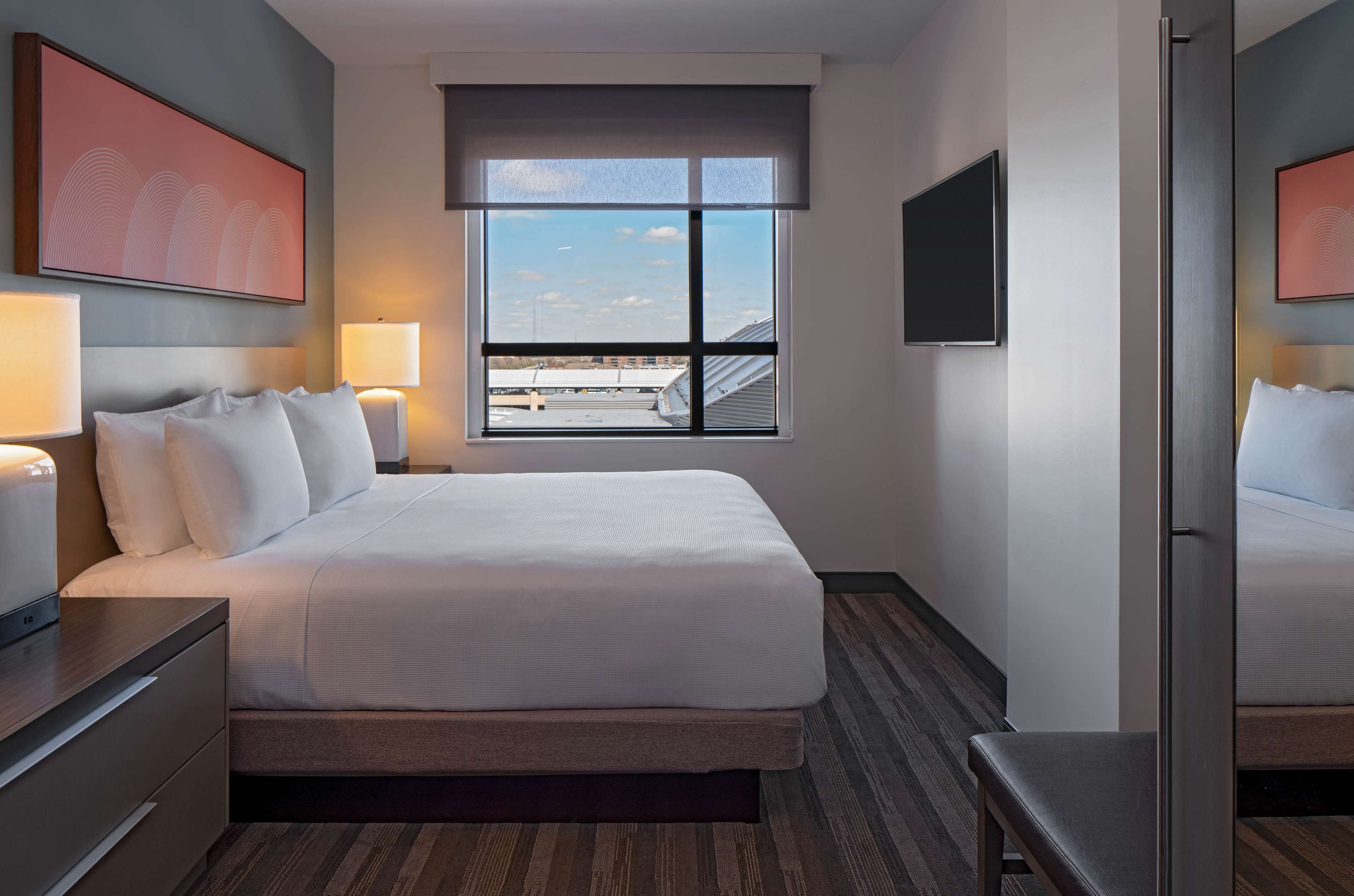 Ein Hotelzimmer mit einem großen Bett, Nachttischlampen, einem an der Wand montierten Fernseher und einem Fenster mit Blick auf Gebäude.