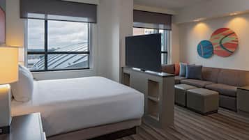 A hotel room with a bed, a TV, a sofa, and a large window with a view of buildings.