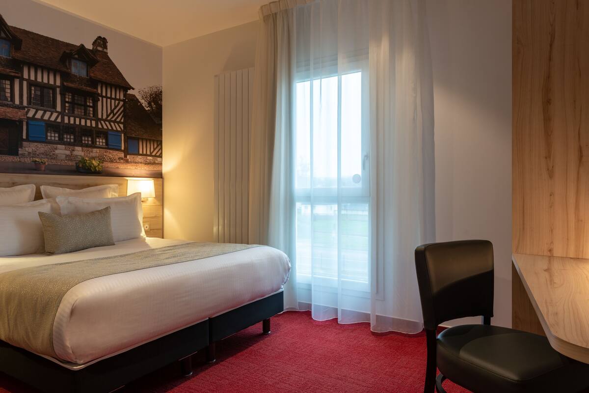 Une chambre d’hôtel avec un grand lit, une chaise, une fenêtre avec des rideaux légers et un mur orné d’une façade de bâtiment de style traditionnel.
