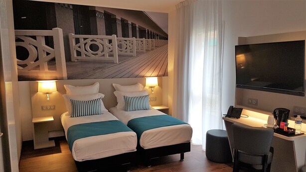 Habitación de hotel con dos camas, un escritorio con silla, un televisor grande y vistas a una terraza de madera a través de cortinas traslúcidas.