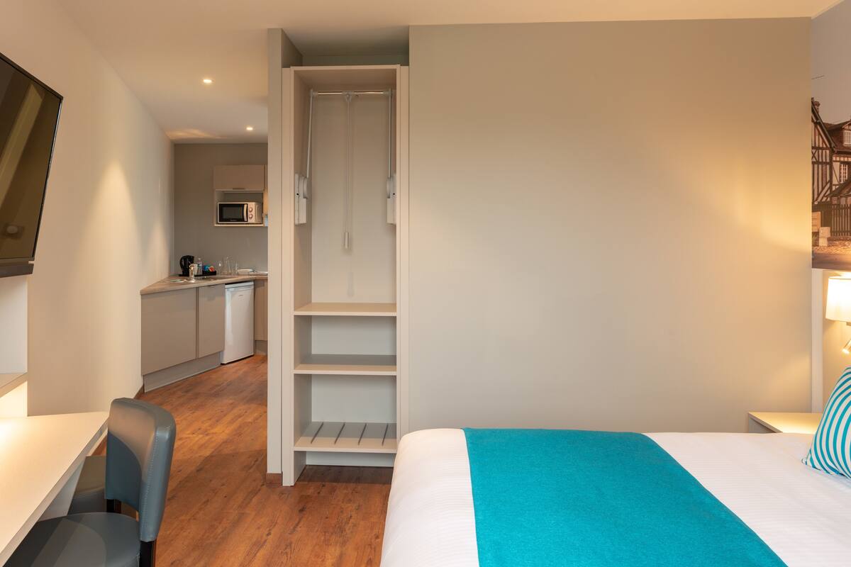 Une chambre d’hôtel de taille réduite, comprenant un lit, un bureau, une chaise, un coin cuisine et une armoire.