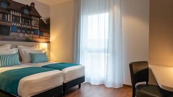 Una habitación de hotel con una cama grande, dos mesitas de noche, una silla y un ventanal con cortinas traslúcidas.
