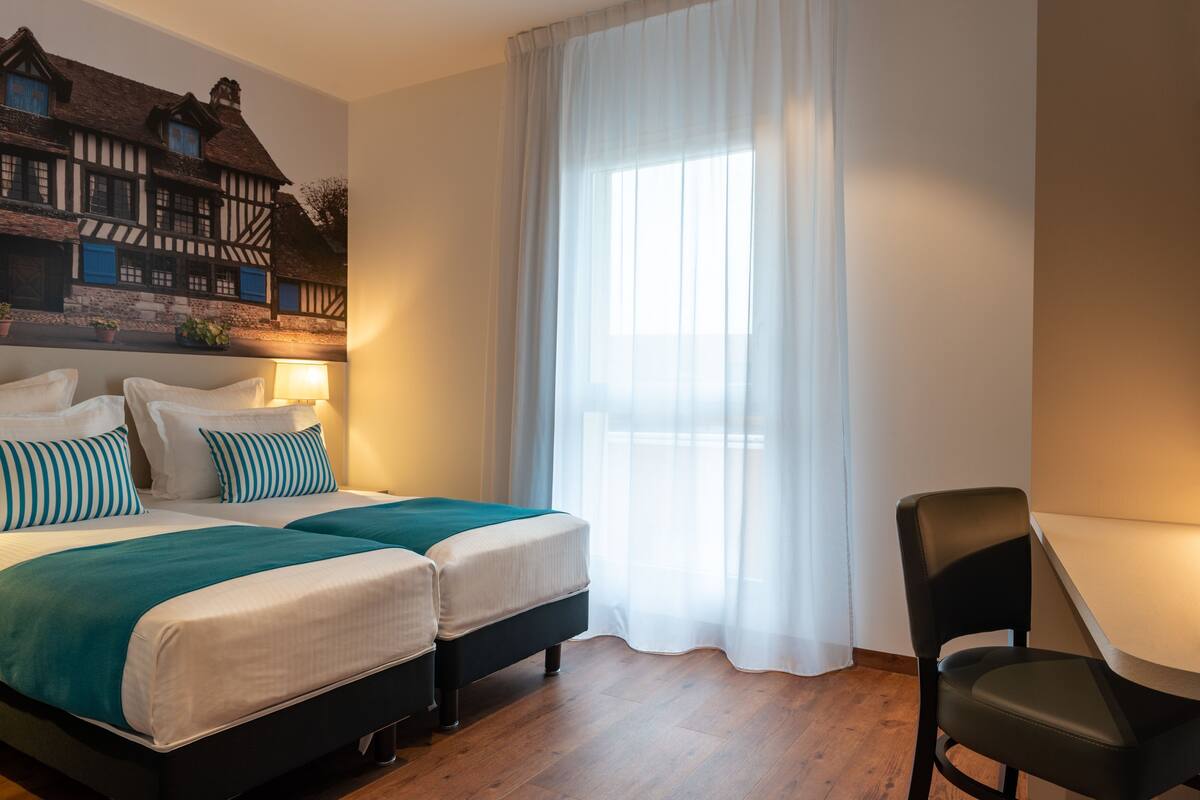 Une chambre avec un lit, deux tables de chevet, une chaise, une grande fenêtre avec des rideaux légers et une fresque murale.