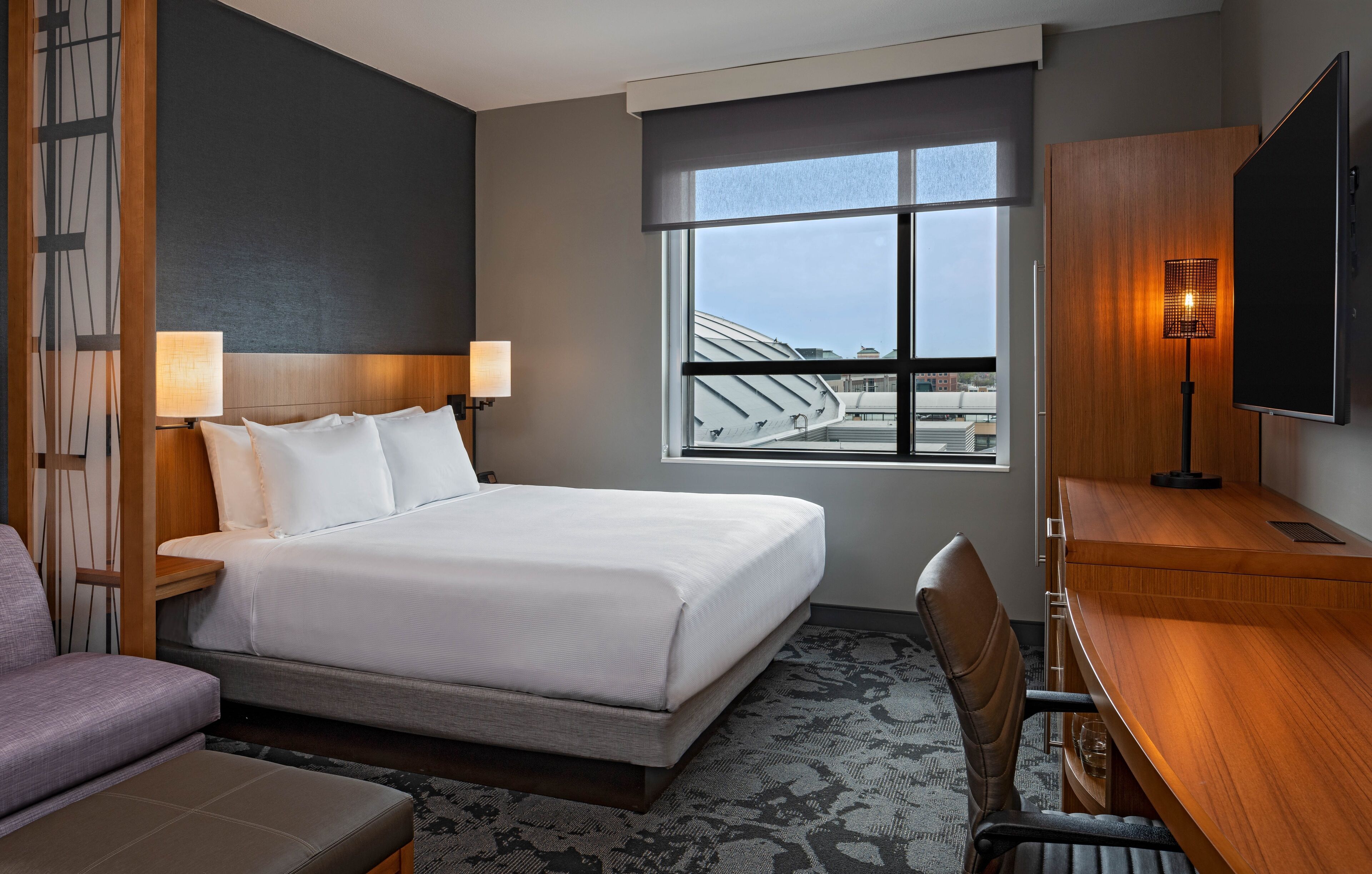 Une chambre d’hôtel avec un grand lit, un bureau, une chaise, une télévision et une fenêtre donnant sur des immeubles.