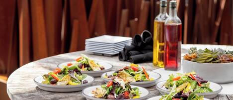 Una mesa de madera redonda con varios platos de ensaladas mixtas de verduras, una pila de servilletas y dos botellas de aceite de oliva.