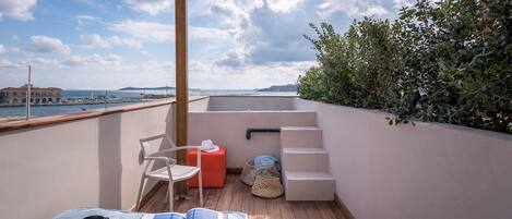 Ein Balkon mit Holzboden, einem weißen Stuhl, einem roten Hocker und einem Tisch mit Tablett, Tassen und einem Buch.