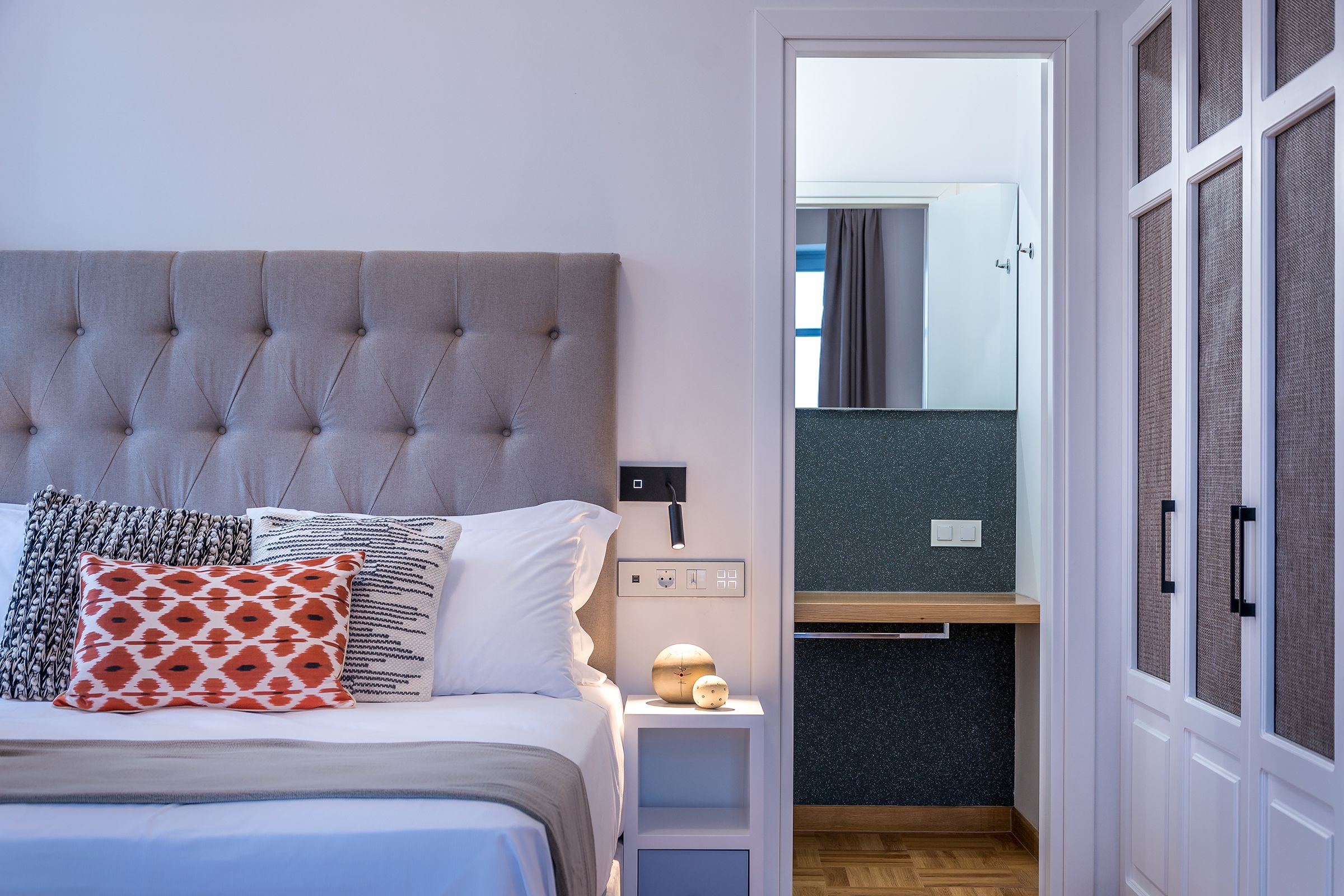 A bedroom with a tufted headboard, a bedside table with a lamp, and a view of a bathroom through an open door.