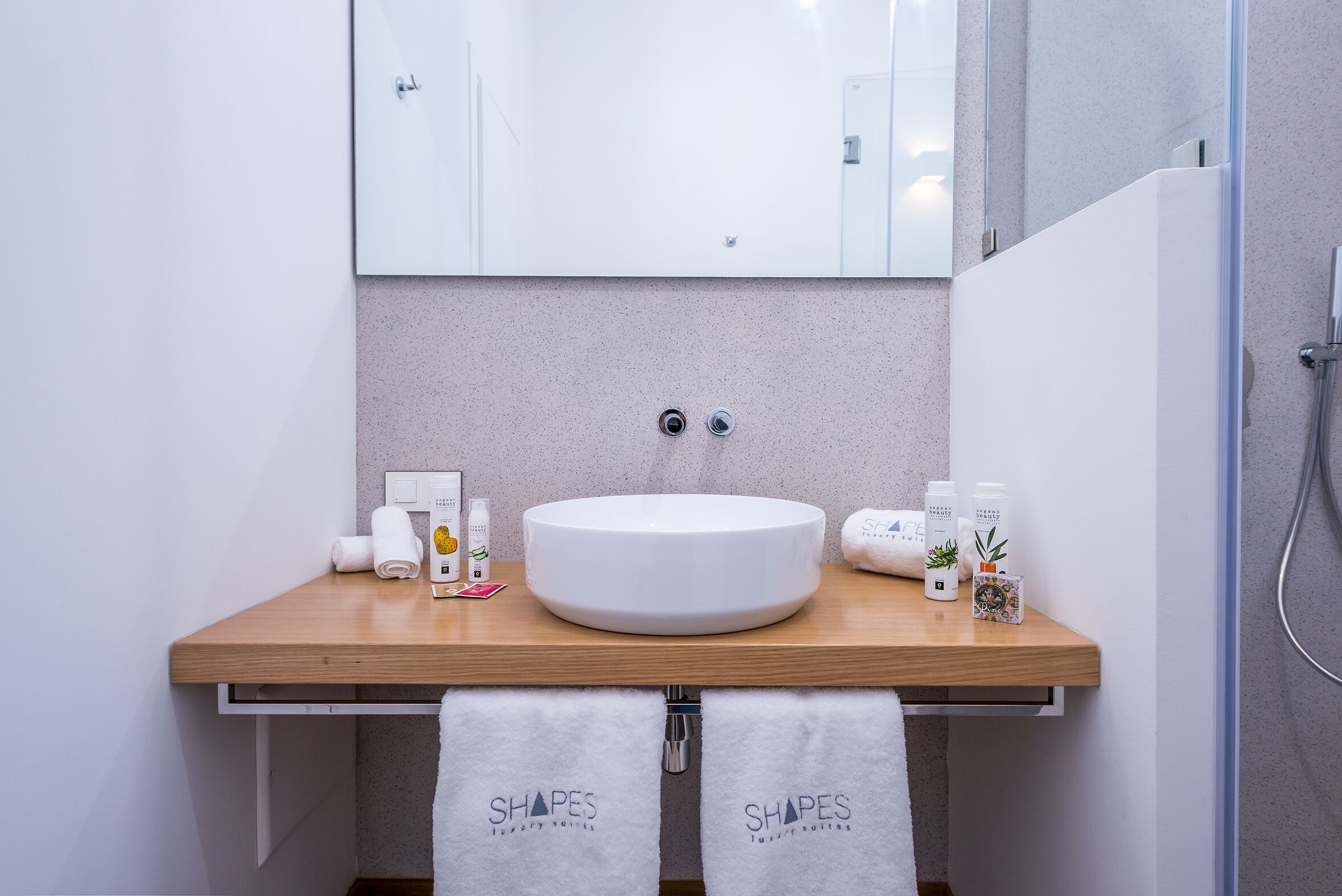 A bathroom with a wooden vanity, a white sink, and a mirror above it. There are towels hanging on the wall and a shower visible to the right.