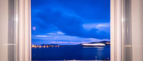A view through a window of a coastal area at dusk with a calm sea, distant city lights, and a clear sky.