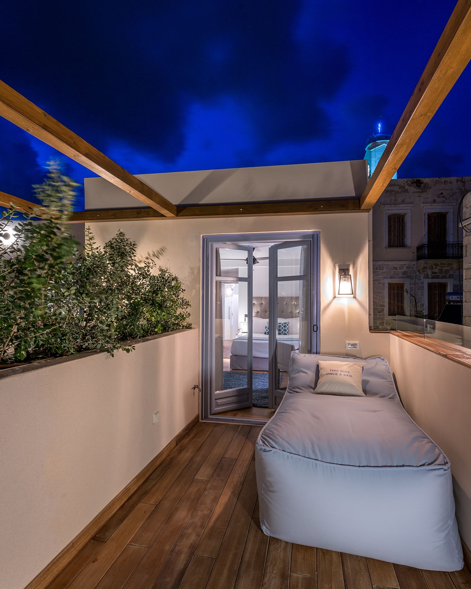 A cozy balcony with a wooden floor, a comfortable bed, and a view of the city at night.