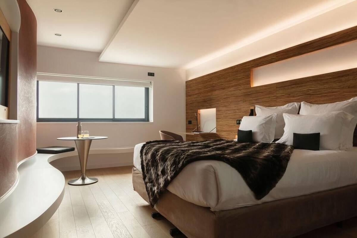 Una habitación de hotel moderna con una cama grande, una mesita redonda pequeña, una silla y un ventanal amplio.