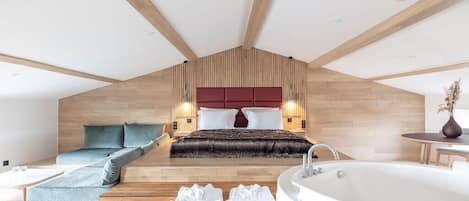 Un dormitorio moderno con techo inclinado, una cama grande con cabecera roja, una zona de estar con sofá, una bañera y suelo de madera.