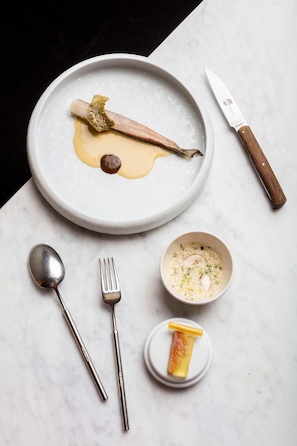 A plate with a piece of fish, a spoon, a fork, and a knife on a marble surface. - Origines (Le Broc)