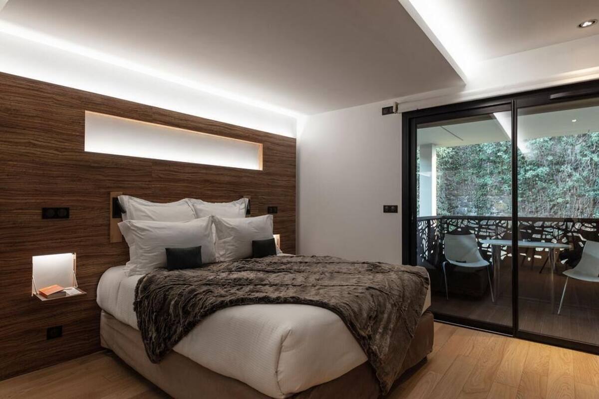 Un dormitorio moderno con cabecera de madera, una cama grande con ropa de cama blanca, una mesita de noche con lámpara y un balcón con mobiliario exterior.