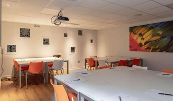 Un salón de conferencias con mesas blancas y sillas naranja, un proyector en el techo y arte abstracto en las paredes.