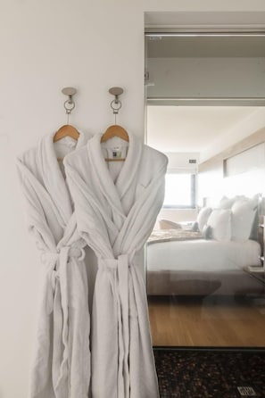 Two white bathrobes hanging on a wall, a bed with white bedding, and a window with daylight. - Origines (Le Broc)
