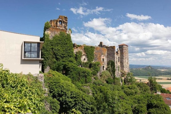 A historic castle ruin with a modern building adjacent to it, surrounded by lush greenery. - Origines (Le Broc)