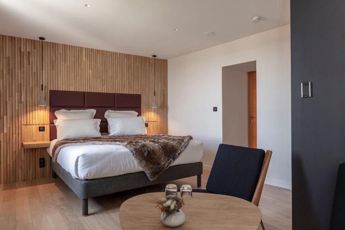 A modern bedroom with a large bed, bedside lamps, a round table with a vase, and a wooden wall paneling.