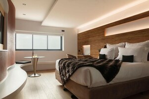 Una habitación de hotel moderna con una cama grande, una mesita redonda pequeña, una silla y un ventanal amplio.