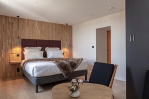 A modern bedroom with a large bed, bedside lamps, a round table with a vase, and a wooden wall paneling. - Origines (Le Broc)