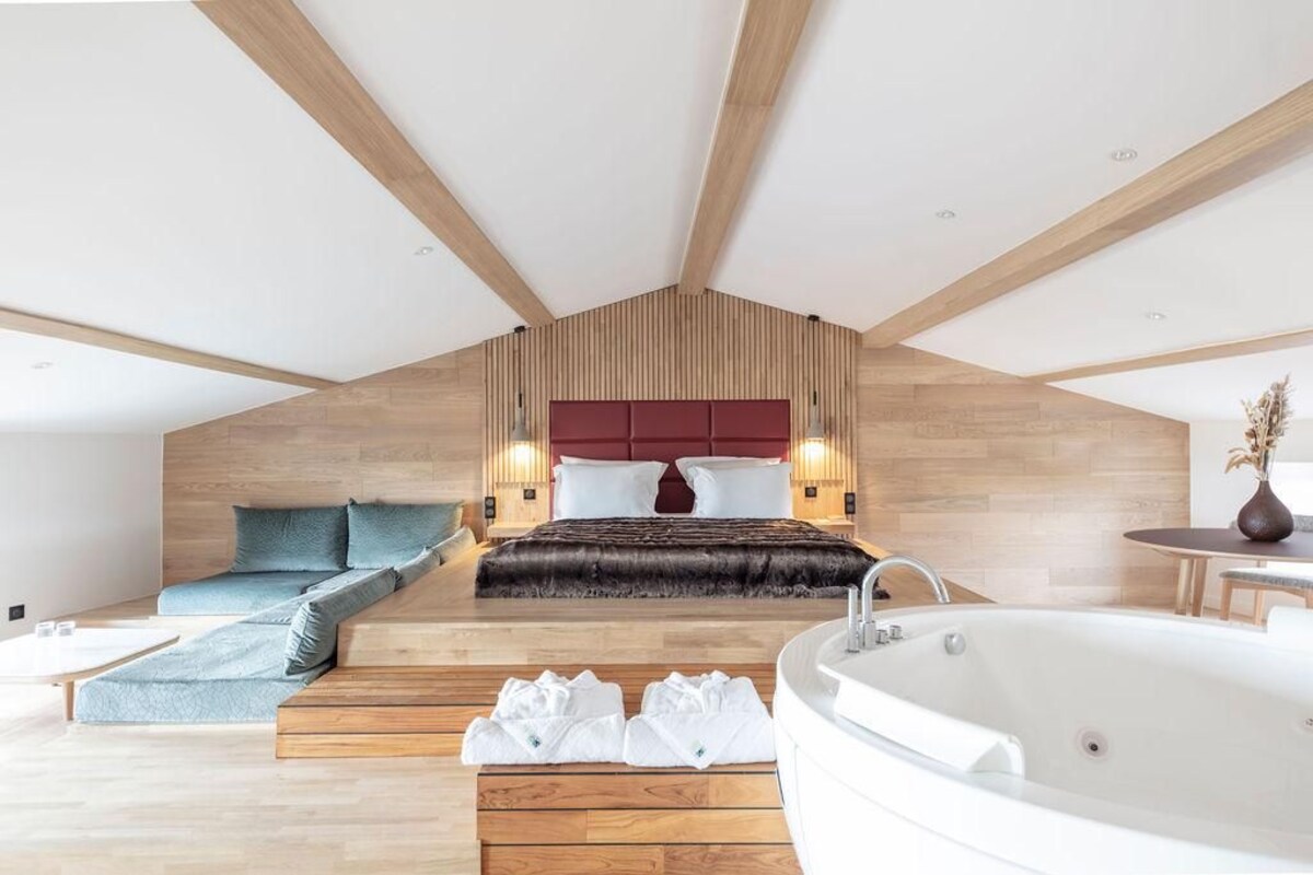 A modern bedroom with a sloped ceiling, a large bed with a red headboard, a sitting area with a sofa, a bathtub, and wooden flooring.