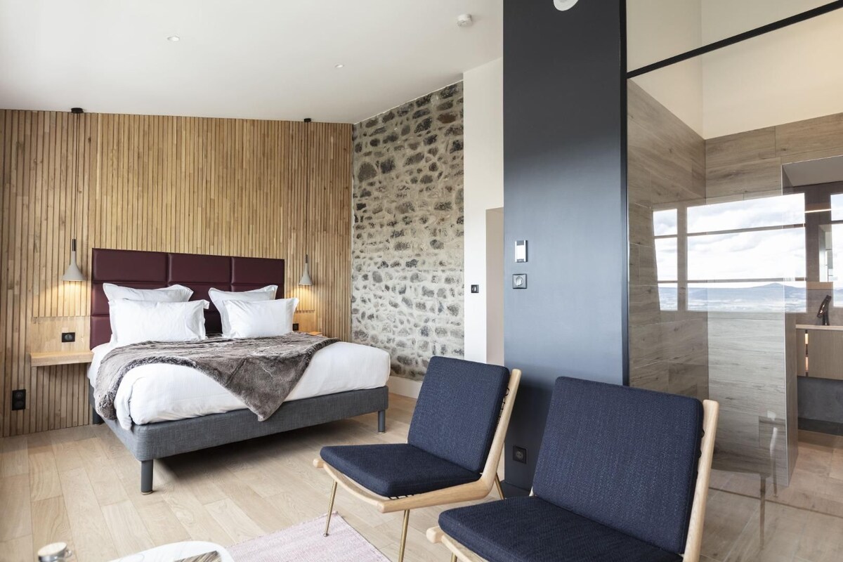 A modern bedroom with a large bed, two bedside tables, a stone accent wall, and a view of the outdoors.