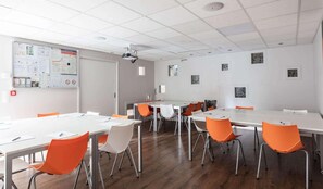 Un salón de conferencias con mesas blancas y sillas anaranjadas, una pizarra con gráficos y un proyector en el techo.