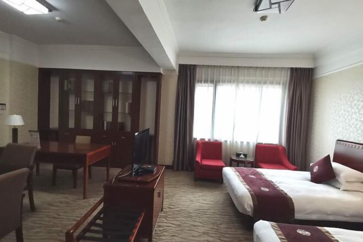 A hotel room with a bed, two red chairs, a desk with a TV, and a window with curtains.