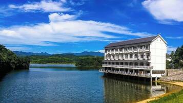 A multi-story building with a flat roof, situated on stilts over a body of water, with a clear sky and mountains in the background.