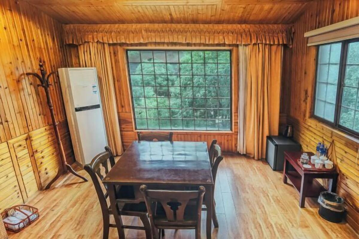 A wooden cabin interior with a dining table, chairs, a refrigerator, a window with curtains, and a small table with cups and bottles.