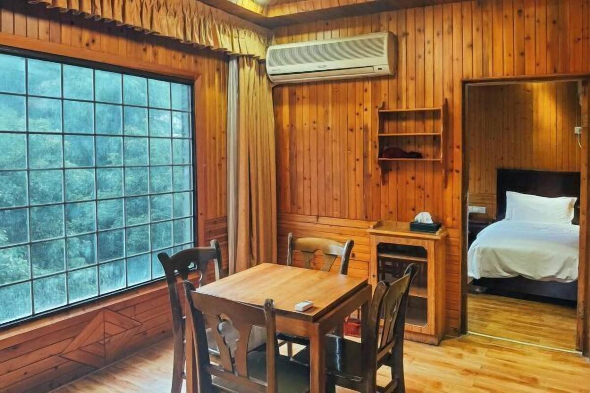 A cozy cabin room with a wooden interior, a dining table with chairs, a bed, and a large window offering a view of trees.