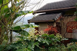 Un edificio japonés tradicional con tejado de tejas, rodeado de exuberante follaje y plantas en flor.