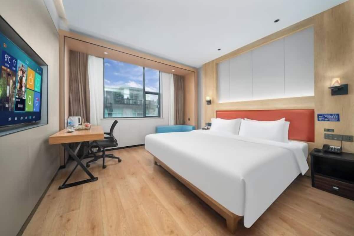 A hotel room with a large bed, a desk with a chair, a TV mounted on the wall, and a window with a view of buildings.